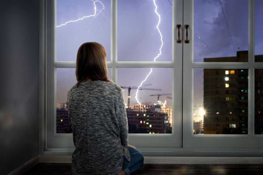 Woman looking out window at storm after losing power and learning how to get your home generator running quickly and safely.